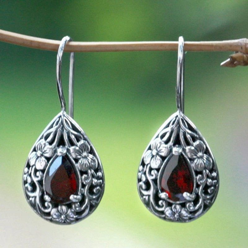 Pirate teardrop earrings with deep red garnet-colored stones and intricate silver metalwork, hanging from a branch.
