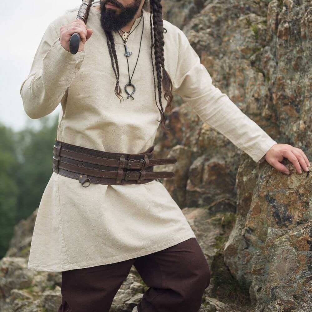 Viking-style man in a natural linen Viking tunic, brown pants, braided hair, layered belt, and runic necklaces.