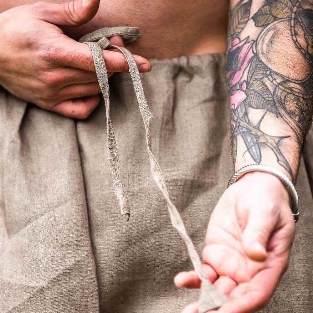 Close-up of hands tying the drawstring waist of stone grey linen Pirate pants.