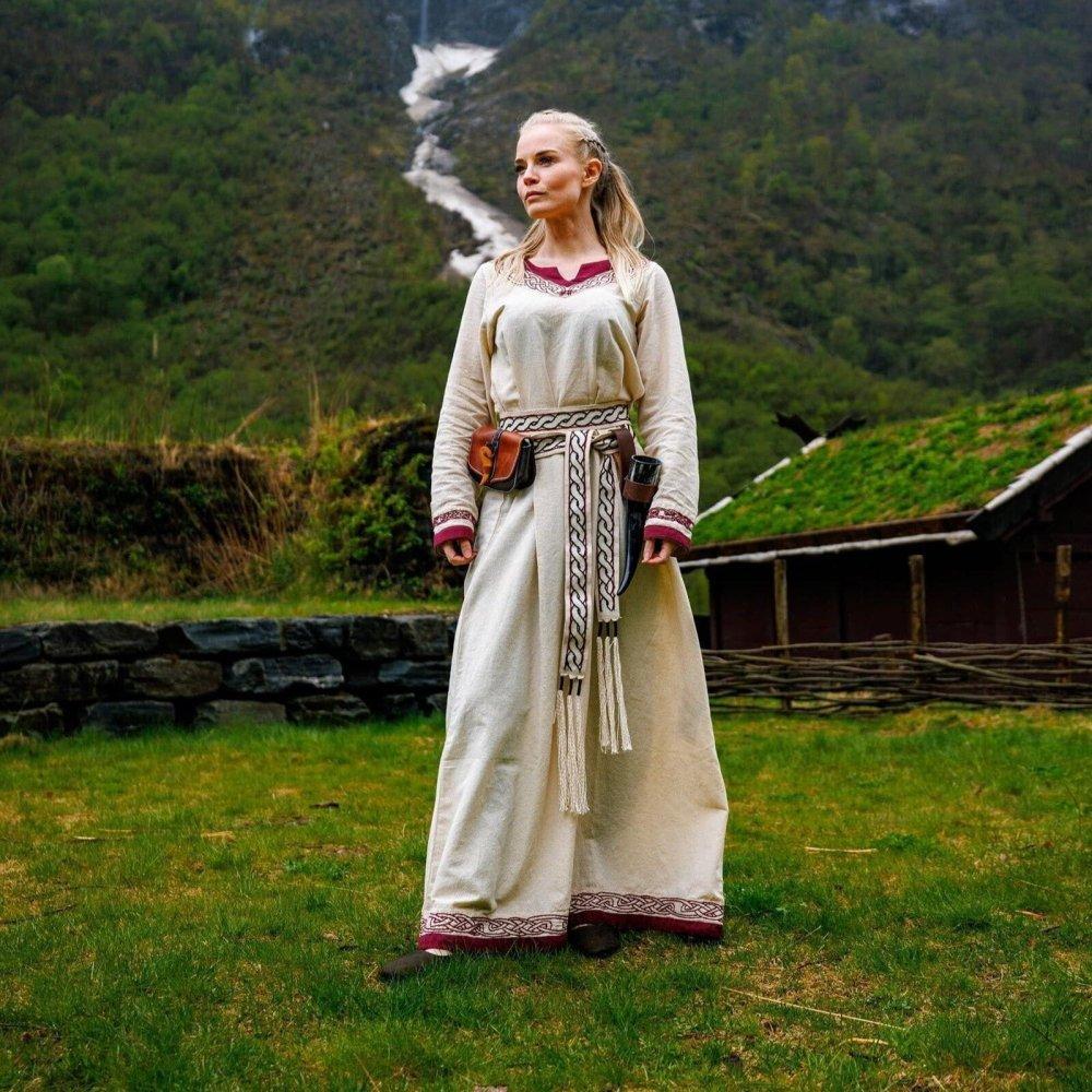 Pirate woman in a cream-colored dress with a woven tie belt, standing in a grassy field.
