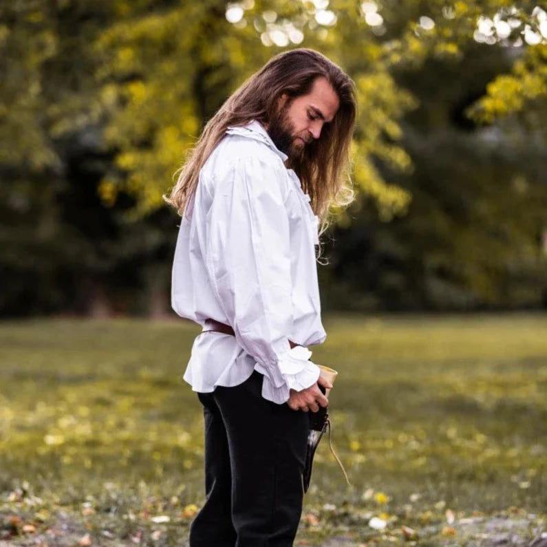 Man in a white cotton Pirate shirt with ruffles, puffed sleeves, and a brown belt, standing outdoors.