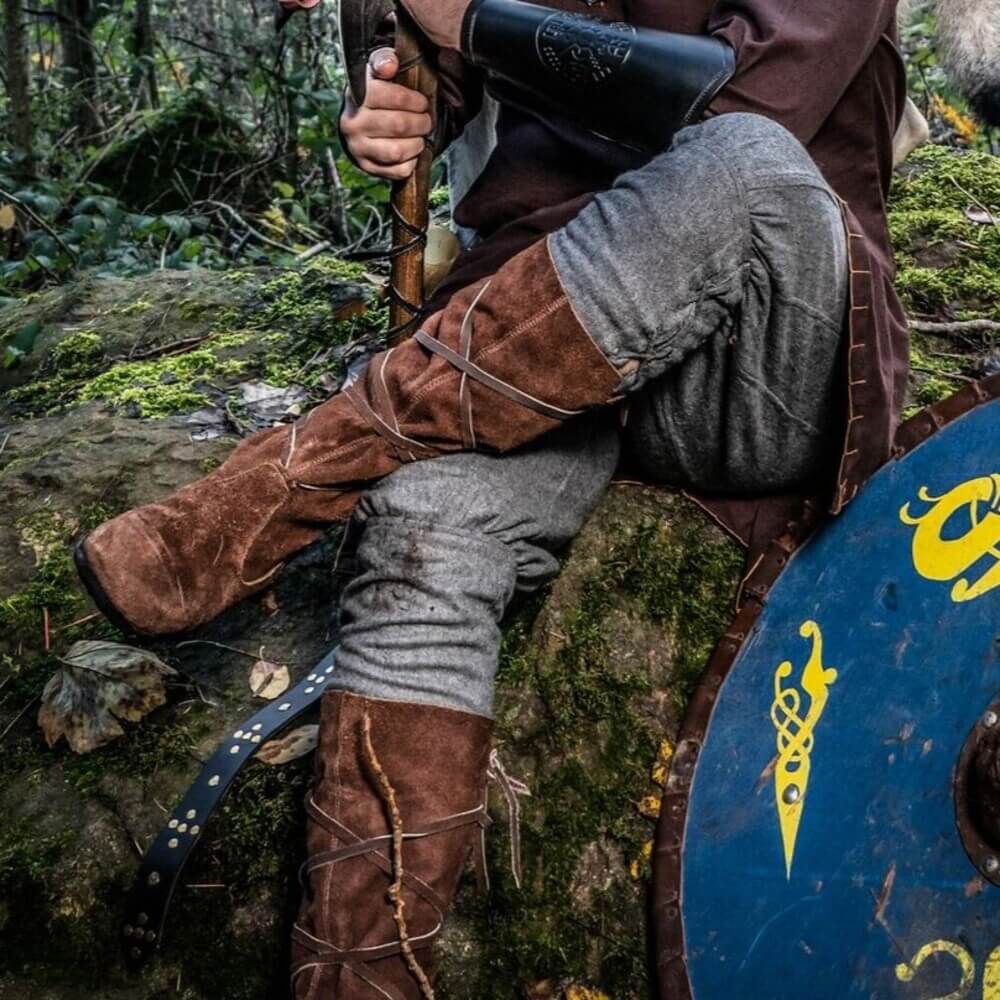 Person wearing brown suede Pirate wrap-tied boots, seated on mossy rock beside a blue Norse shield in a forest.