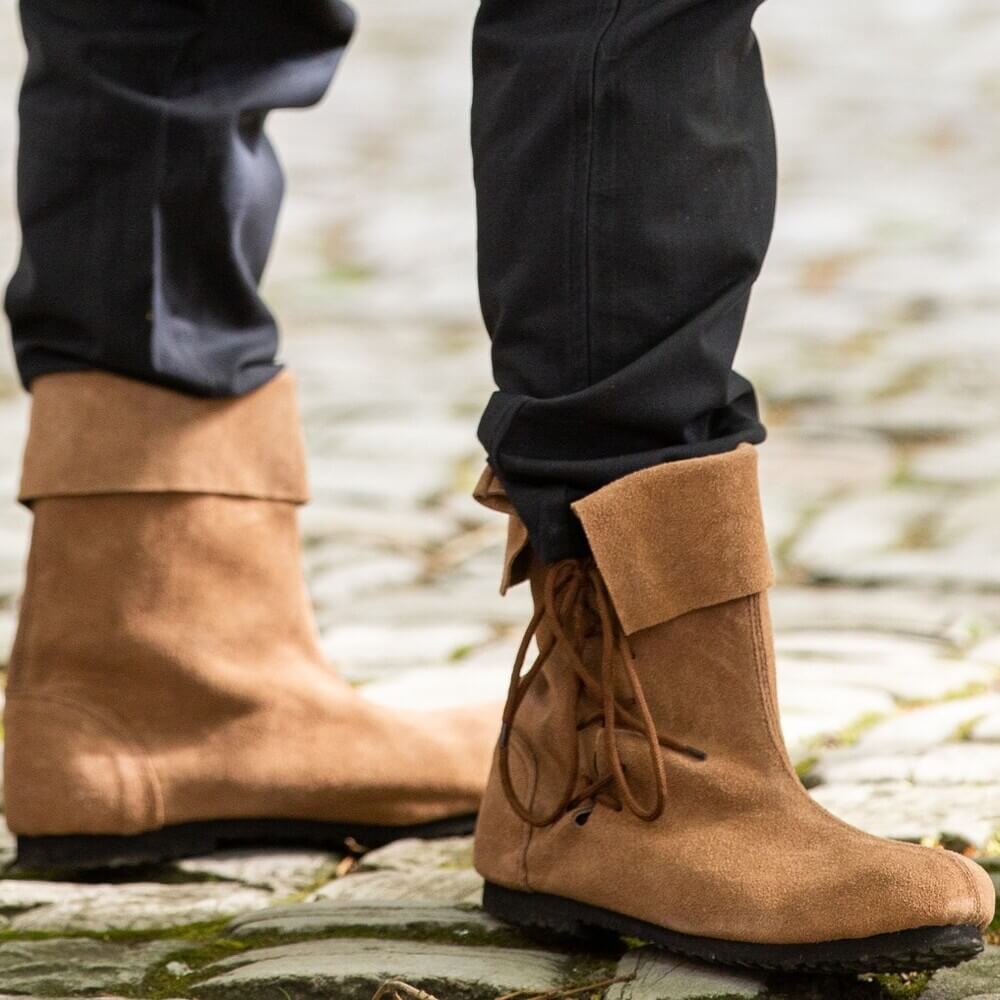 Person standing on cobblestones wearing Pirate Sigurd brown suede boots with soft fold-over tops and side laces.