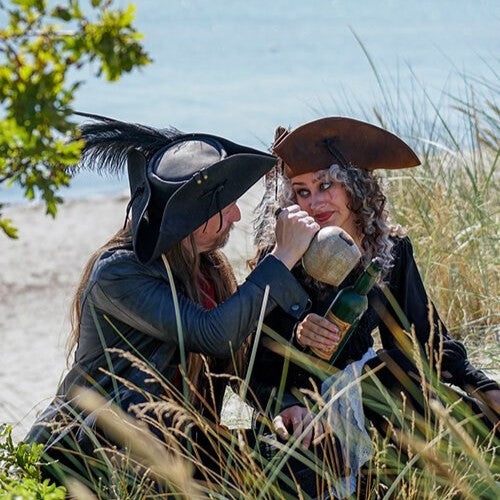 Two people in pirate costumes by the beach; one wears a leather tricorn hat and holds a bottle.