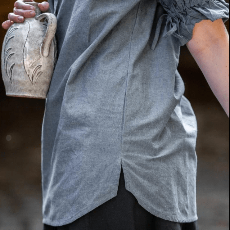 Grey-blue Pirate blouse, hand-woven cotton, short sleeve, straight cut with side slits, worn over dark pants.