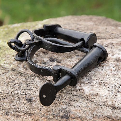 Black wrought iron Pirate manacles with hand-riveted cuffs and a key-turned lock, resting on a rock.