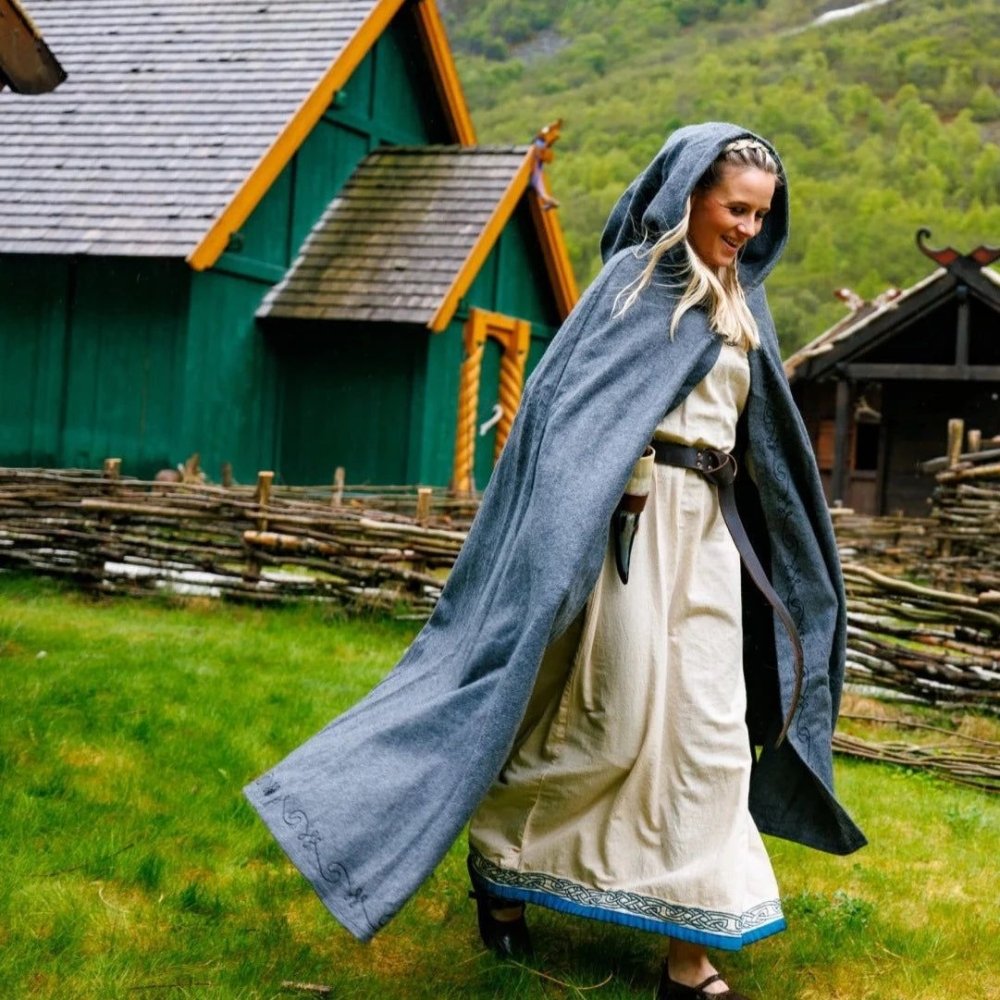Woman in a long, grey wool Pirate cloak with hood and embroidered trim, walking outdoors.