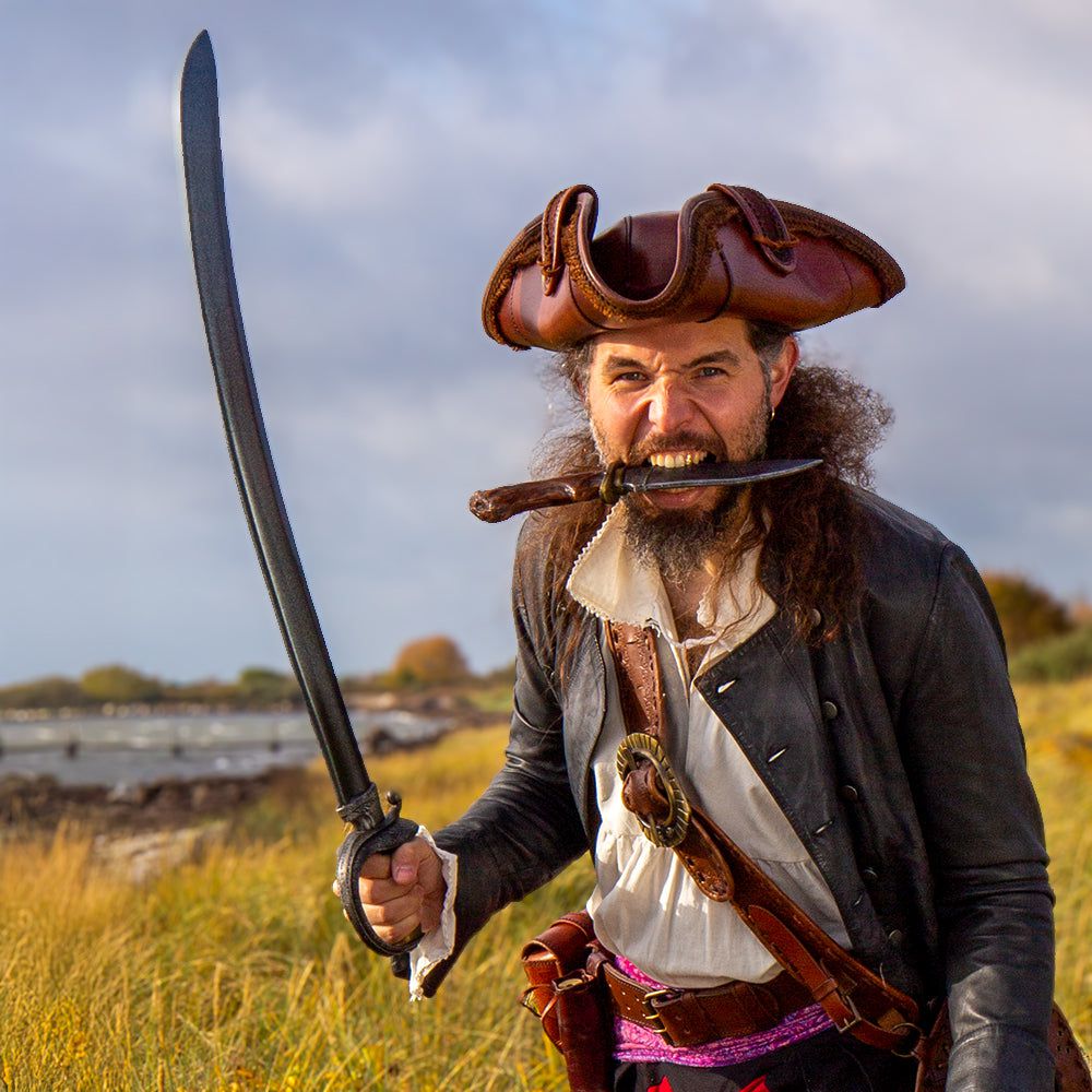 A fierce Pirate in a brown hat and black jacket holds a dark curved cutlass, a dagger in his mouth.