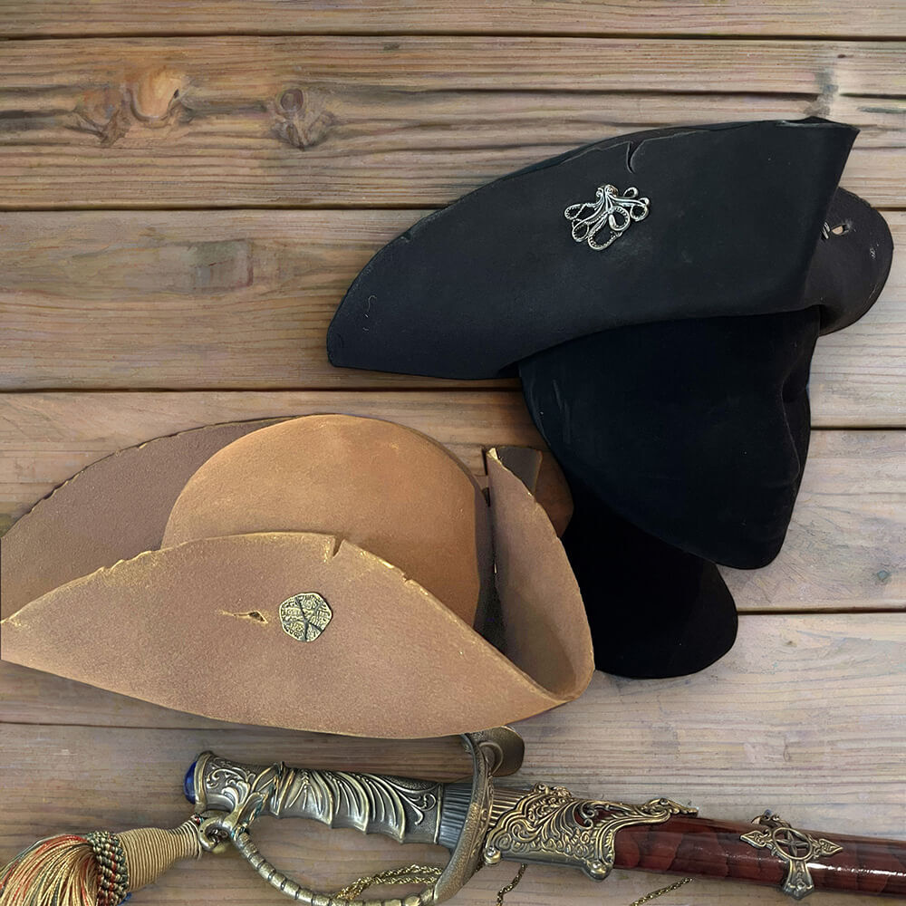 Two weathered felted wool Pirate tricorn hats, one black with a silver kraken pin, one tan with a gold coin pin, beside a sword.