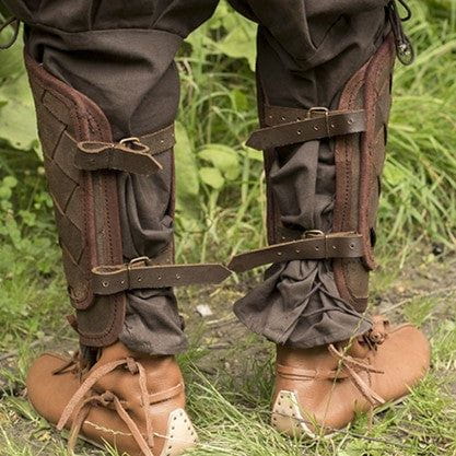 Brown woven leather Pirate greaves with brass buckles worn over dark pants and light brown shoes.