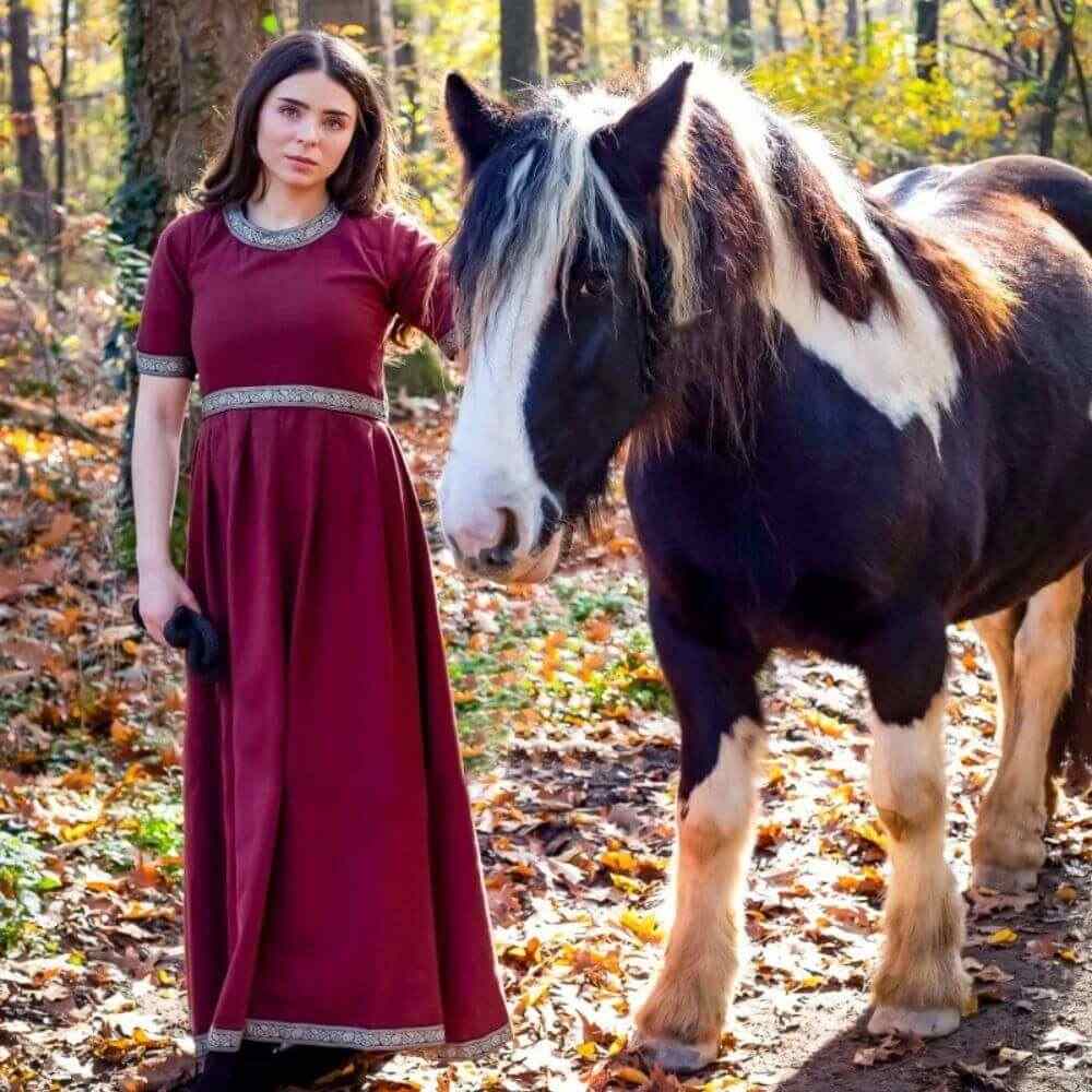 Woman in a red short sleeve pirate dress standing in autumn woods beside a draft horse